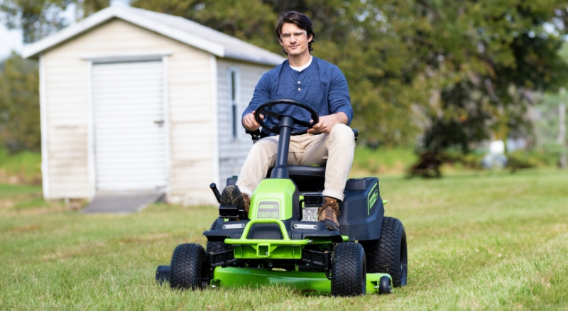 Homeowner mows grass using riding mower with white shed and trees in background